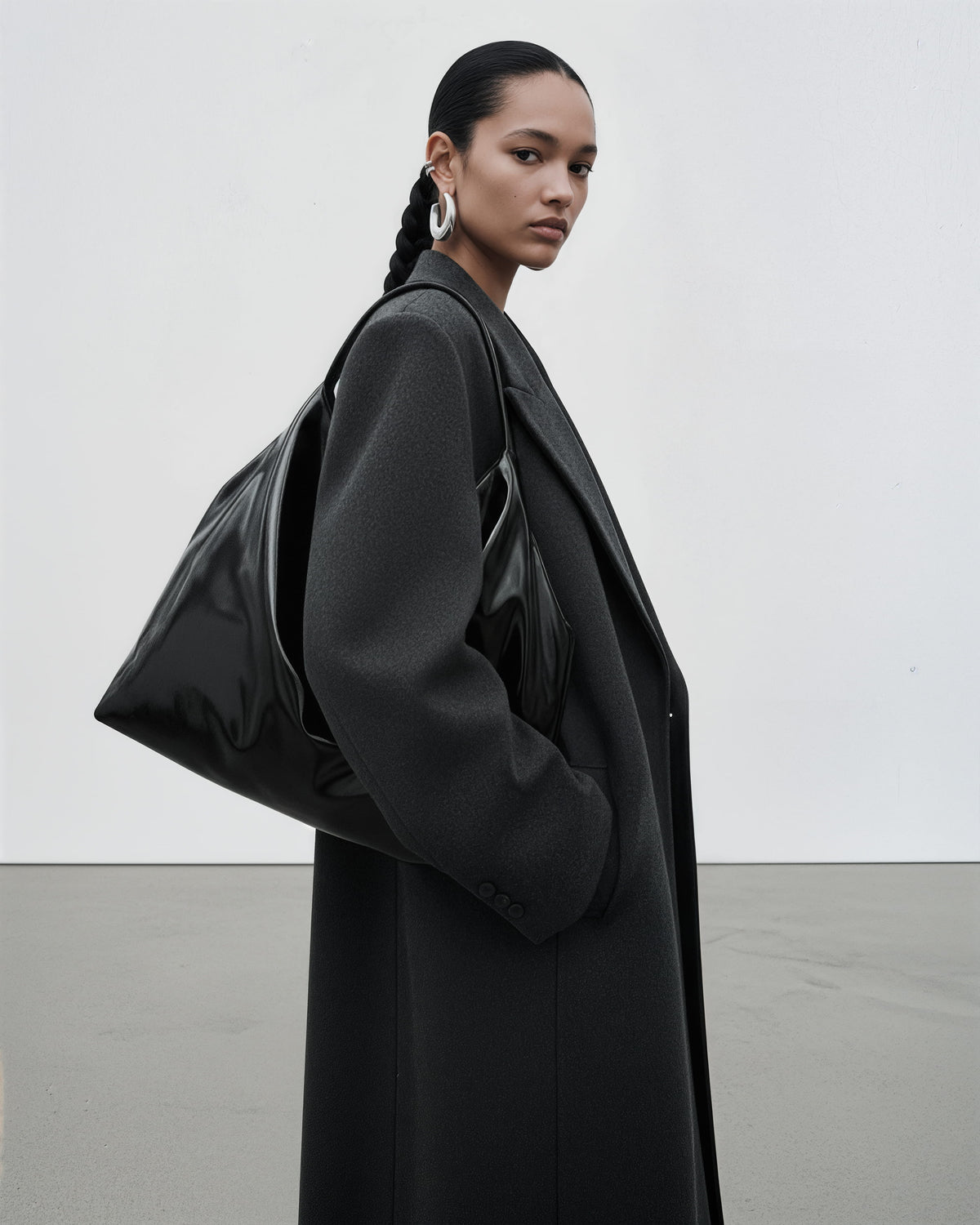 A model in a black coat and black hair bra carries a large black shoulder bag with a glossy finish, set against a minimalist light gray studio background.
